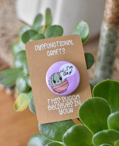 lilac purple button badge has a smiling little succulent, speech bubble coming from near the mouth says "hi I'm gay" 